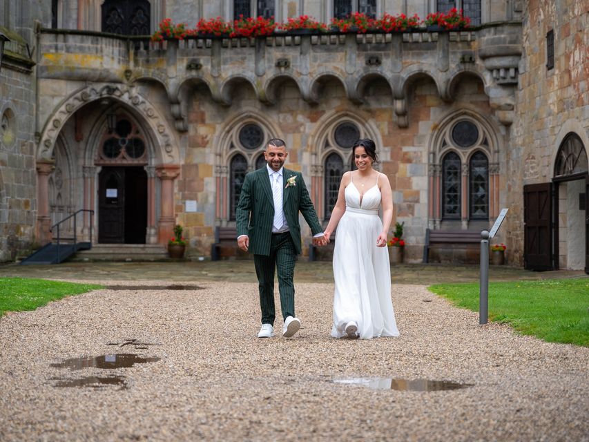 Ein Brautpaar läuft Händchen haltend auf einem Weg vor einem Schloss. Die Braut trägt ein weisses Kleid mit Schleier, der Bräutigam einen eleganten Anzug. Das Schloss im Hintergrund ist eindrucksvoll und verleiht der Szene eine romantische und märchenhafte Atmosphäre.
