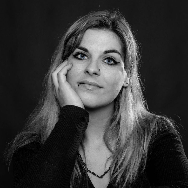 Ein Frauenportrait in Schwarz-Weiss. Die Frau schaut ausdrucksstark in die Kamera, wobei Licht und Schatten ihre Gesichtszüge betonen. Der Hintergrund ist neutral gehalten, sodass der Fokus auf ihrem Gesicht liegt.