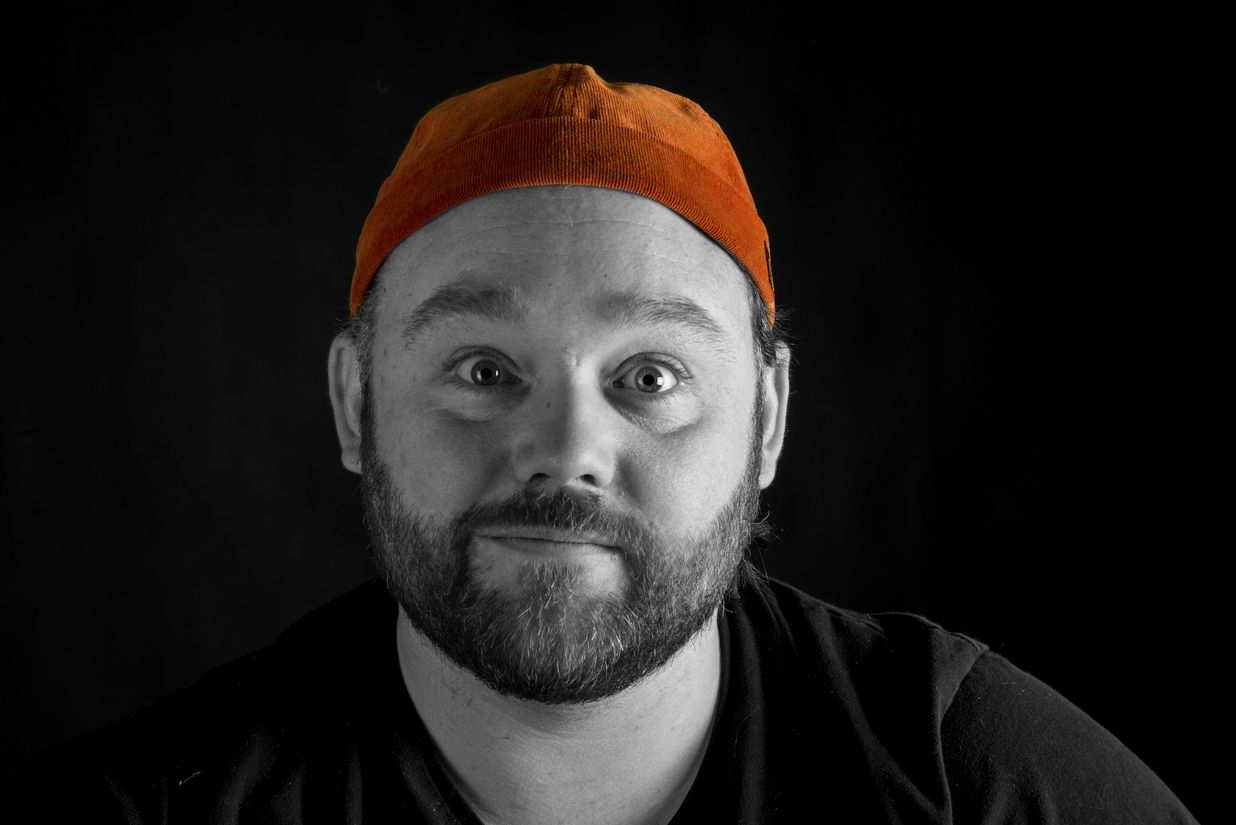 Ein Mann schaut direkt in die Kamera. Das Bild ist in Schwarz-Weiss gehalten, wobei nur seine orangene Mütze farbig hervorsticht. Der Kontrast zwischen der monochromen Darstellung und der leuchtenden Mütze lenkt den Blick auf das auffällige Accessoire.