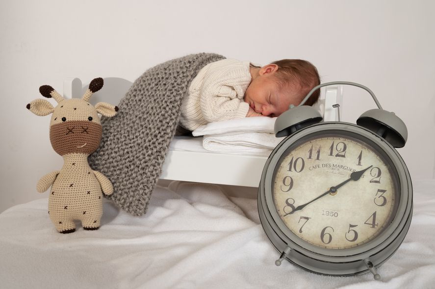 Ein Säugling liegt schlafend in einem kleinen Bett, zugedeckt mit einer weichen Decke. Vor dem Bett steht eine Uhr mit klar sichtbarem Zifferblatt.