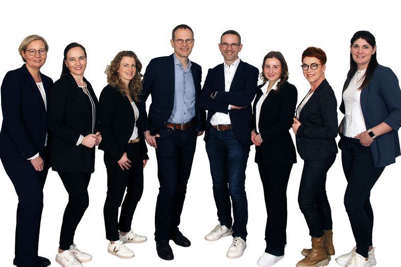 Ein Gruppenfoto mit 8 Personen, die zusammen in einem professionellen Umfeld stehen. Die Personen sind geschäftlich gekleidet und lächeln in die Kamera. Der Hintergrund deutet auf ein Büro oder einen neutralen Firmenkontext hin.